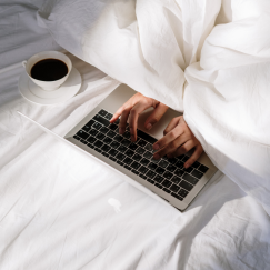 Hands typing on laptop in bed with coffee